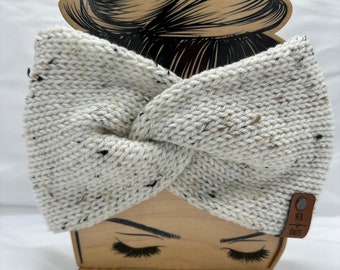 May include: White knitted headband with a speckled pattern. The headband is tied in a knot and is displayed on a wooden mannequin head. The headband has a brown leather tag with the text "MTB Knits" on it.