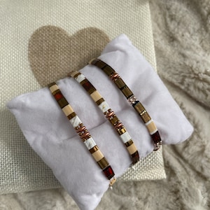 May include: Three beaded bracelets with a mix of colors, including brown, cream, and red, are displayed on a white pillow. The bracelets have a variety of bead shapes and sizes. A small, cream-colored bag with a heart design is in the background.