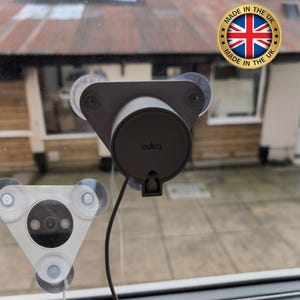 May include: A black and grey security camera with a suction cup mount attached to a window. The camera has a label that says "ooloo" on it. The camera is connected to a wire that runs to a white triangular device with a lens and three suction cups.