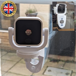 May include: A white security camera mounted on a window using a suction cup. The camera has a black lens and a white casing. The camera is mounted on a white bracket. The camera is facing forward.