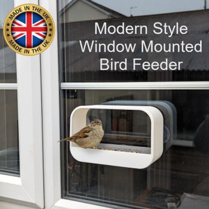 May include: A white window mounted bird feeder with a sparrow perched on the edge. The feeder has a clear front panel and the text "WWW.GGPLUSBD.COM" is printed on the bottom. The feeder is attached to a window with suction cups.