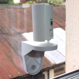 May include: White security camera mounted on a white window mount. The camera is attached to a window with a suction cup.