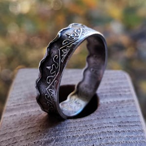 May include: A silver ring with an intricate, etched design. The ring features a wavy edge and detailed patterns, including floral and geometric shapes. The ring is displayed on a wooden surface.