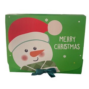 Puede incluir: Caja de regalo verde con un mu&ntilde;eco de nieve que lleva un gorro de Pap&aacute; Noel rojo y una bufanda verde. La caja tiene un lazo verde y dice "Feliz Navidad" en la parte delantera.