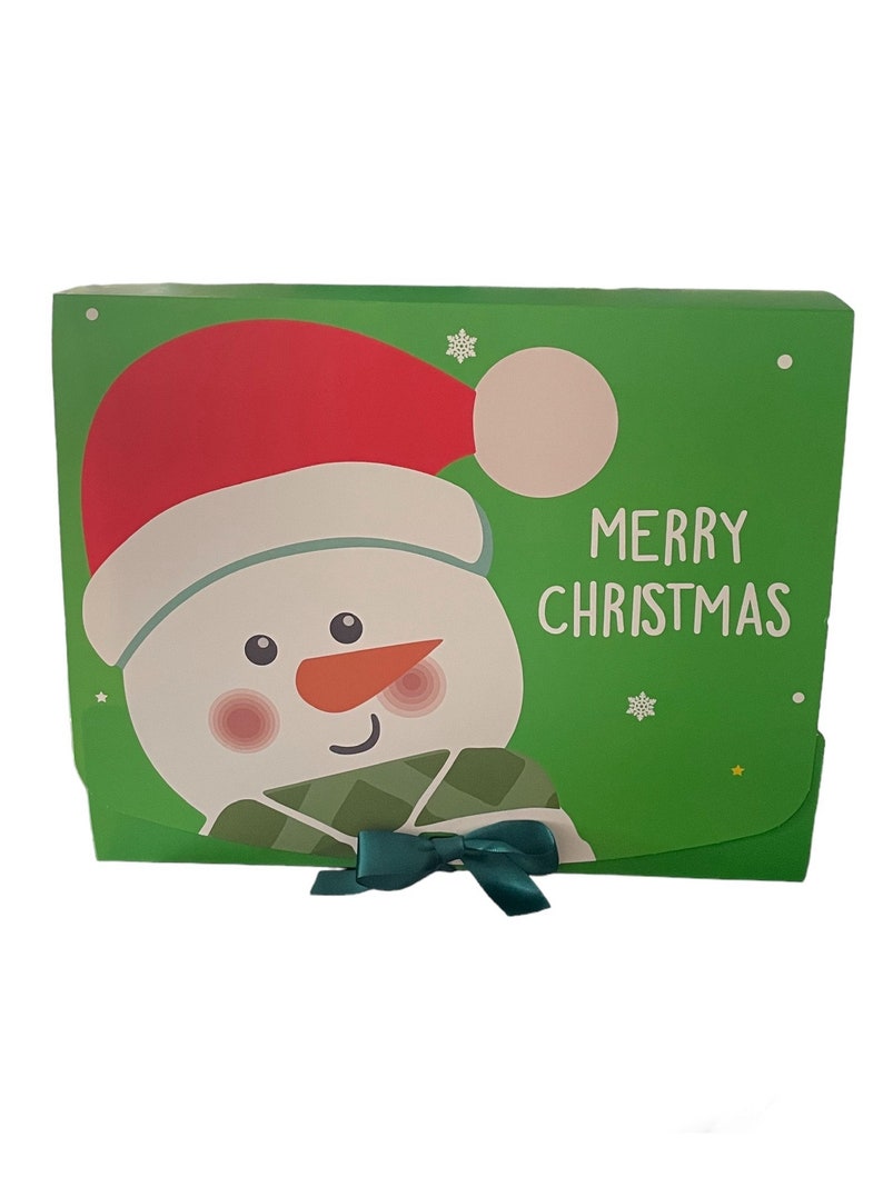 Puede incluir: Caja de regalo verde con un mu&ntilde;eco de nieve que lleva un gorro de Pap&aacute; Noel rojo y una bufanda verde. La caja tiene un lazo verde y dice "Feliz Navidad" en la parte delantera.