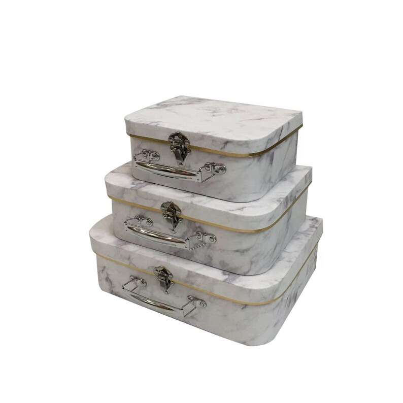 May include: A stack of three decorative storage boxes with a marble pattern. Each box has a silver handle and clasp, with a gold-colored trim. The boxes are rectangular with rounded corners and are arranged in a descending size order.