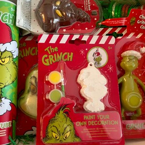 Puede incluir: Un conjunto de regalo de Navidad con tem&aacute;tica de Grinch con un personaje verde de Grinch en un tubo rojo y blanco, un adorno blanco de Grinch para pintar, una figura verde de Grinch y una moneda de chocolate de Grinch.