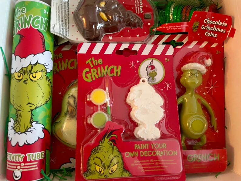Puede incluir: Un conjunto de regalo de Navidad con tem&aacute;tica de Grinch con un personaje verde de Grinch en un tubo rojo y blanco, un adorno blanco de Grinch para pintar, una figura verde de Grinch y una moneda de chocolate de Grinch.