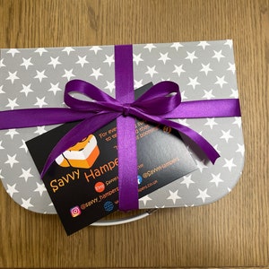 May include: A gift box with a gray star pattern, tied with a purple ribbon and bow. A black card with the text "Savvy Hampers" and social media handles is attached to the ribbon. The box is on a wooden surface.