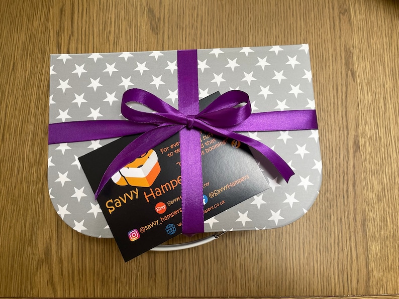 May include: A gift box with a gray star pattern, tied with a purple ribbon and bow. A black card with the text "Savvy Hampers" and social media handles is attached to the ribbon. The box is on a wooden surface.
