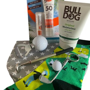 May include: A gift set with golf-themed items, including a green sock, sunscreen, lip balm, and moisturiser. It features a golf ball, a golf club keyring, and a golf ball marker. The box has a star pattern.
