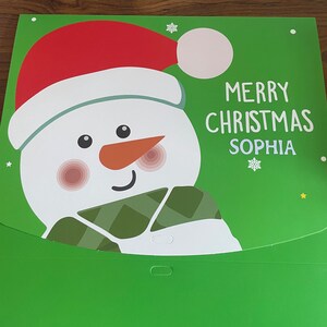 Puede incluir: Una tarjeta de Navidad verde con un mu&ntilde;eco de nieve que lleva un gorro de Pap&aacute; Noel rojo y una bufanda verde. La tarjeta dice "Feliz Navidad Sophia".