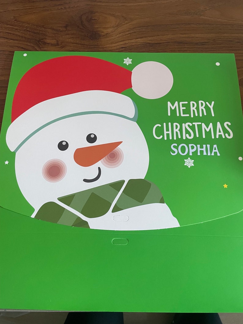 Puede incluir: Una tarjeta de Navidad verde con un mu&ntilde;eco de nieve que lleva un gorro de Pap&aacute; Noel rojo y una bufanda verde. La tarjeta dice "Feliz Navidad Sophia".