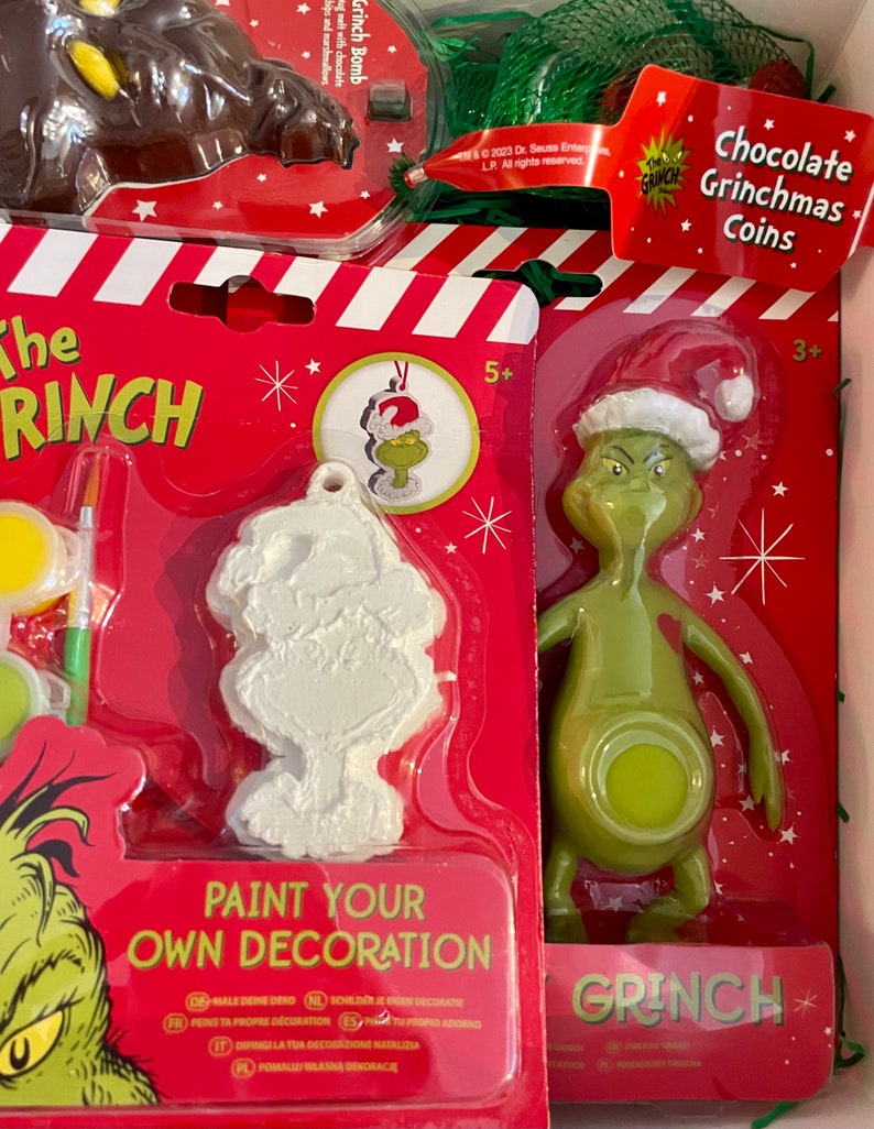 Puede incluir: Un kit de manualidades de Navidad con el Grinch. El kit incluye un adorno de Grinch para pintar, una figura de Grinch y monedas de chocolate Grinchmas. El embalaje es rojo y blanco con las palabras "The Grinch" y "Pinta tu propia decoraci&oacute;n".