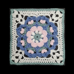 May include: A crocheted square featuring a floral design in shades of pink, blue and green. The centre of the square has a large flower with a smaller flower in the centre.