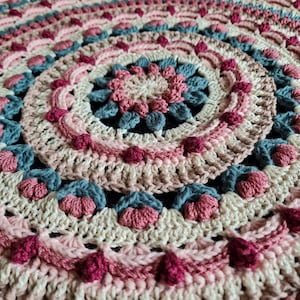May include: A crocheted round rug with a circular pattern in shades of pink, beige, and blue. The rug has a textured surface with raised stitches and a fringe border.