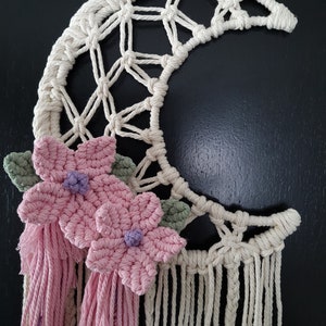 Peut inclure: Suspension murale en macramé en forme de croissant de lune crème avec des fleurs roses, des feuilles vertes et des pompons roses. La lune a un motif tissé et une frange de cordons noués. Les fleurs ont des centres violets.