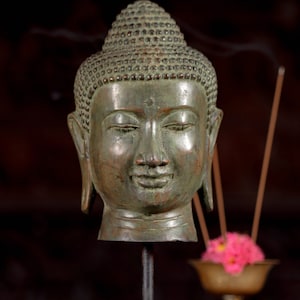 May include: A green bronze Buddha head sculpture on a black stand. The sculpture has a serene expression and a detailed head.