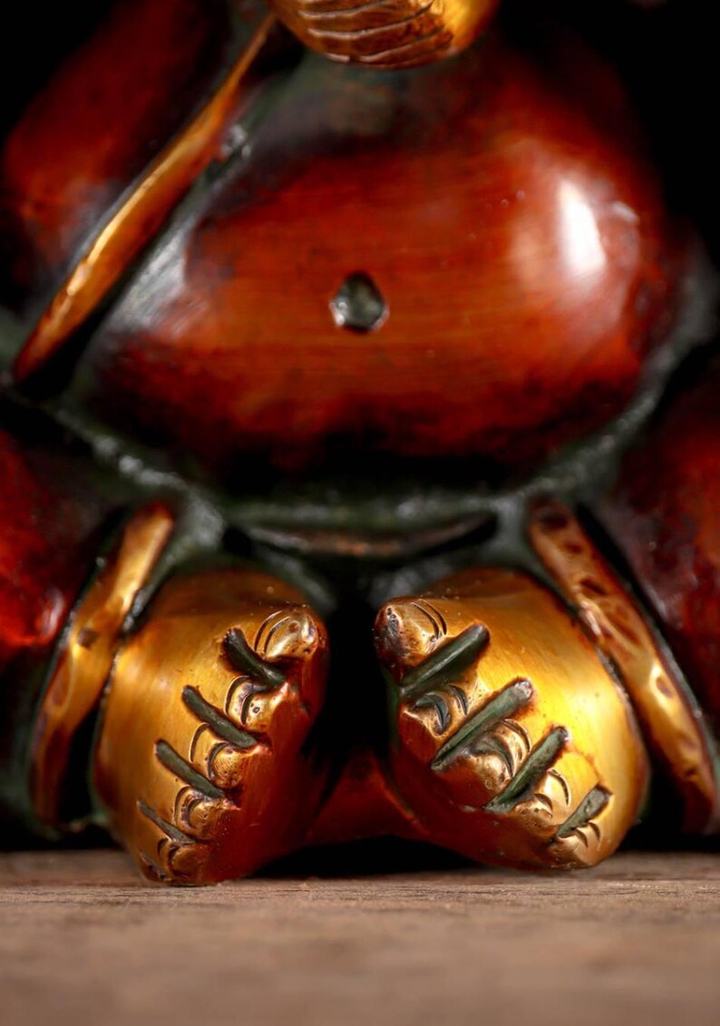 Puede incluir: Primer plano de una estatua dorada de Ganesha con intrincados detalles en las manos y los pies. La estatua est&aacute; hecha de lat&oacute;n y tiene una p&aacute;tina marr&oacute;n rojiza.