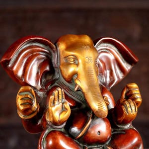 Puede incluir: Una estatua de bronce dorada y roja de Ganesha, el dios hind&uacute; de los comienzos, la sabidur&iacute;a y la prosperidad. La estatua representa a Ganesha sentado con la trompa levantada y las manos en un gesto mudra. La estatua est&aacute; intrincadamente detallada y tiene un dise&ntilde;o tradicional.