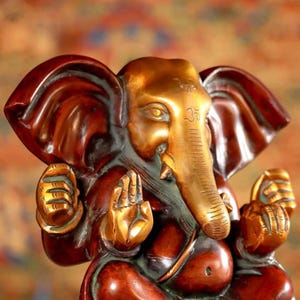 Puede incluir: Una peque&ntilde;a estatua de Ganesha de color dorado y marr&oacute;n con la trompa apuntando hacia la derecha. La estatua est&aacute; sentada con las manos en posici&oacute;n de oraci&oacute;n. La estatua tiene un s&iacute;mbolo Om rojo y dorado en la frente.