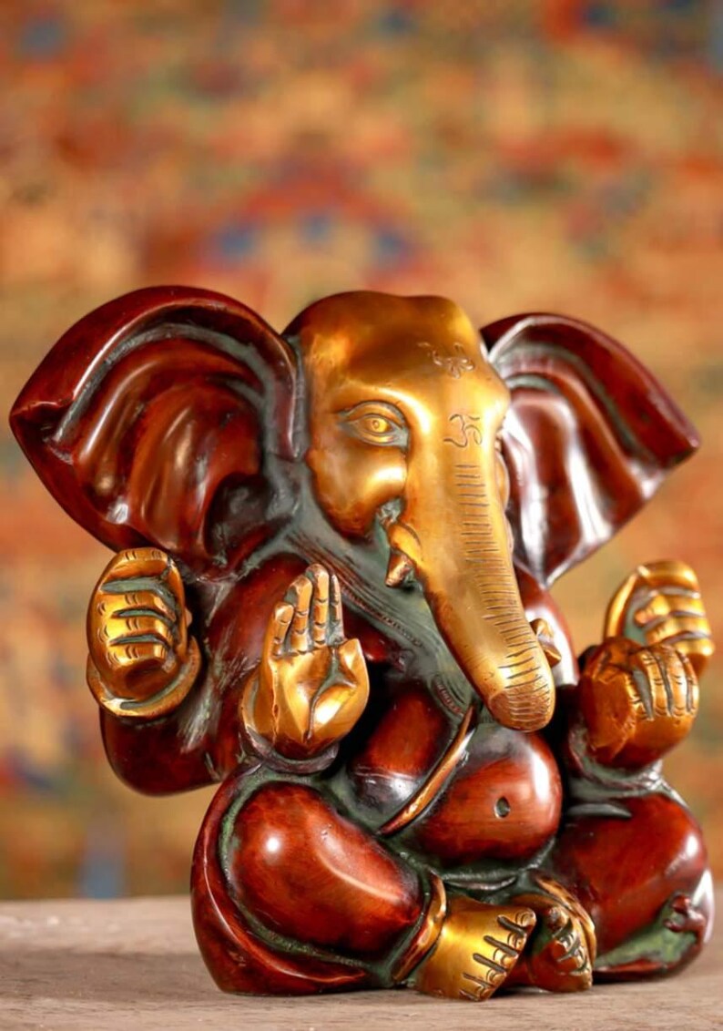 Puede incluir: Una peque&ntilde;a estatua de Ganesha de color dorado y marr&oacute;n con la trompa apuntando hacia la derecha. La estatua est&aacute; sentada con las manos en posici&oacute;n de oraci&oacute;n. La estatua tiene un s&iacute;mbolo Om rojo y dorado en la frente.