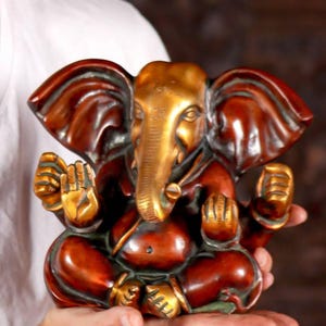 Puede incluir: Una peque&ntilde;a estatua de Ganesha roja y dorada con la trompa enroscada. La estatua est&aacute; sentada en una postura meditativa con las manos en mudra.