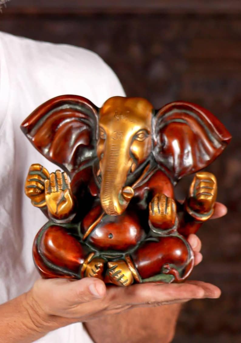 Puede incluir: Una peque&ntilde;a estatua de Ganesha roja y dorada con la trompa enroscada. La estatua est&aacute; sentada en una postura meditativa con las manos en mudra.