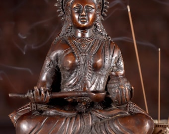 Könnte beinhalten: Eine Bronzestatue einer sitzenden Göttin mit einer Krone und filigranen Details. Die Statue hält eine kleine Schale und einen langen, dünnen Gegenstand in ihren Händen. Die Statue steht auf einem Holzsockel mit einem geschnitzten Design. Die Statue ist von Rauch aus Räucherstäbchen umgeben.