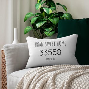 May include: White rectangular cushion with the text "HOME SWEET HOME" above the numbers "33558" and "TAMPA, FL" in black. The cushion is on a grey sofa with a cream-coloured blanket. A green plant and a dark green cushion are in the background.