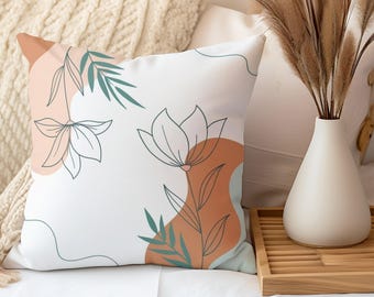 Könnte beinhalten: Ein weißes Kissen mit einem minimalistischen Blumendesign mit grünen Blättern und weißen Blüten auf weißem Hintergrund. Das Kissen hat eine braune und blaue abstrakte Form im Hintergrund.