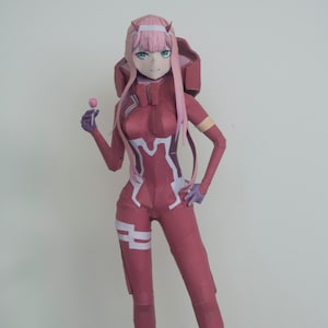 Pode incluir: Uma figura de papelcraft rosa e branca de uma personagem feminina de anime com cabelo longo rosa, vestindo um macacão vermelho e branco. A figura está segurando uma pirulito rosa e está em pé sobre uma base preta com o texto "Darling in the Franxx".
