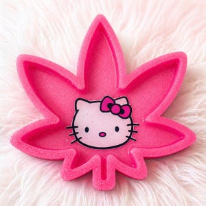 May include: A pink, glittery tray in the shape of a cannabis leaf. The tray features a Hello Kitty design in the center. The tray is a bright pink color and sits on a white, fluffy surface.