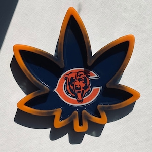May include: A navy blue, leaf-shaped ashtray with an orange border. The center features the Chicago Bears logo in orange and white. The ashtray is made of a glossy material and sits on a white surface.