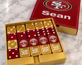 Puede incluir: Un juego de dominó en una caja dorada con una tapa a juego que muestra el logo de los San Francisco 49ers y el nombre Sean. Las fichas son rojas y doradas, algunas con puntos blancos, otras con el logo de los 49ers y el nombre Sean en blanco.