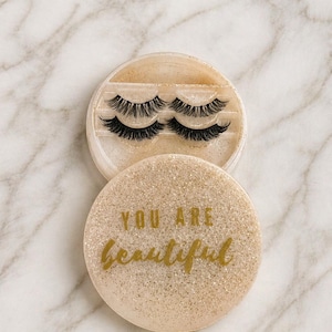May include: A set of false eyelashes in a round, glittery case. The case is open, revealing two sets of black lashes. The bottom of the case has the words "YOU ARE beautiful" in gold script.