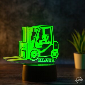 May include: Green illuminated 3D acrylic night light shaped like a forklift with the name "KLAUS" underneath. The night light is on a black base and is sitting on a wooden surface.