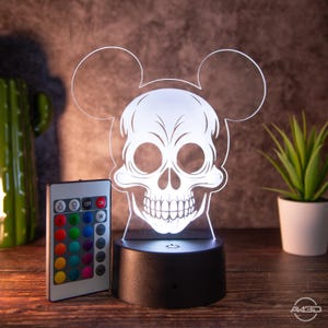 Puede incluir: Un cráneo blanco con orejas de Mickey Mouse se ilumina con una luz nocturna LED 3D. La luz tiene una base negra y viene con un mando a distancia para cambiar el color de la luz.