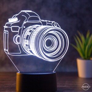 Könnte beinhalten: Eine weiße, 3D-gedruckte Nachtischlampe in Form einer Kamera mit schwarzem Sockel. Die Kamera wird von innen beleuchtet und erzeugt so einen leuchtenden Effekt.