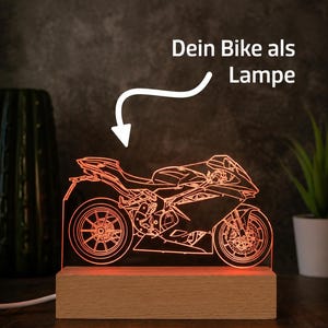 Può includere: Una lampada a LED a luce arancione con una silhouette dettagliata a linee di una moto sportiva montata su una base rettangolare in legno. Sopra la moto, una freccia bianca indica il testo "Dein Bike als Lampe" scritto in bianco.