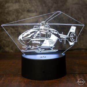 Könnte beinhalten: Ein LED-Hubschrauber-Nachtlicht in Schwarz und Weiß. Das Design zeigt die Texte "Luftrettung", "DRF", "NOTARZT" und "D-HOSTIN". Das Licht steht auf einem schwarzen Sockel und wird auf einer Holzoberfläche angezeigt.