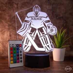 Puede incluir: Una lámpara de hockey 3D impresa en blanco con un mando a distancia. La lámpara está encendida y muestra a un portero de hockey en acción. El portero lleva una máscara y protecciones. La lámpara tiene una base negra. El mando a distancia tiene una variedad de opciones de color.
