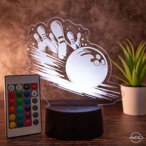 Könnte beinhalten: Ein weißes, 3D-gedrucktes Nachtlicht in Form einer Bowlingkugel und Kegeln. Das Licht befindet sich auf einem schwarzen Sockel und wird mit einer Fernbedienung mit mehreren Farboptionen geliefert.