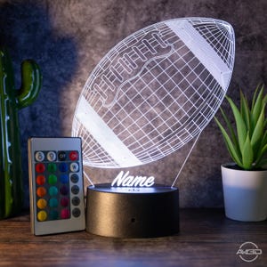 Op de afbeelding: Een wit 3D-geprint nachtlampje in de vorm van een voetbal met een afstandsbediening. De lamp heeft een zwarte basis en de naam "Name" is op de basis gedrukt.