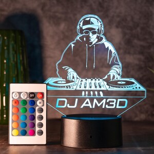 Könnte beinhalten: LED-Nachtlicht mit DJ-Motiv und Fernbedienung. Das Licht zeigt die Silhouette eines DJs mit Kopfhörern und Sonnenbrille, der einen Plattenspieler bedient. Der Sockel ist schwarz, und das Licht strahlt blau. Der Text "DJ AM3D" ist zu sehen.