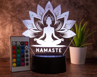 May include: A white 3D printed night light with a silhouette of a person in a lotus pose with the word "NAMASTE" below. The night light is on a black base and has a remote control with multiple color options.