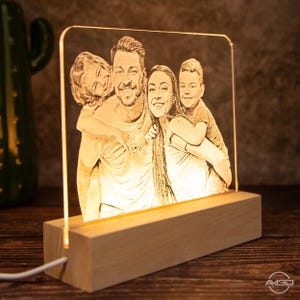 Peut inclure: Une lampe en acrylique personnalis&eacute;e avec une lueur blanc chaud, pr&eacute;sentant une image grav&eacute;e d'une famille de quatre personnes. La lampe repose sur une base en bois.