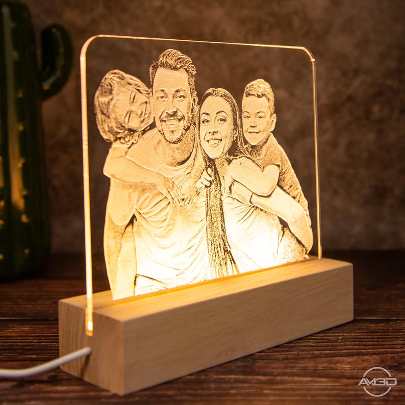 Peut inclure: Une lampe en acrylique personnalis&eacute;e avec une lueur blanc chaud, pr&eacute;sentant une image grav&eacute;e d'une famille de quatre personnes. La lampe repose sur une base en bois.