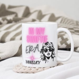 May include: White ceramic mug featuring a pink graphic that reads "In My Swiftie Era" and a black and white illustration of a woman wearing sunglasses. The text "Yanelky" is printed below the graphic.