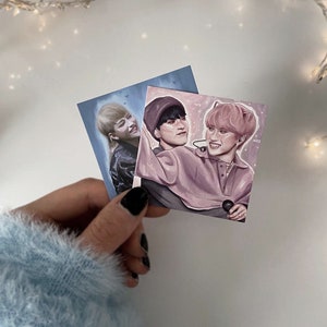 Könnte beinhalten: Zwei quadratische Kunstdrucke mit Porträts von Personen. Ein Druck zeigt eine Person mit blonden Haaren, der andere zwei Personen. Die Drucke werden von einer Hand gehalten, die einen hellblauen Pullover und schwarzen Nagellack trägt.